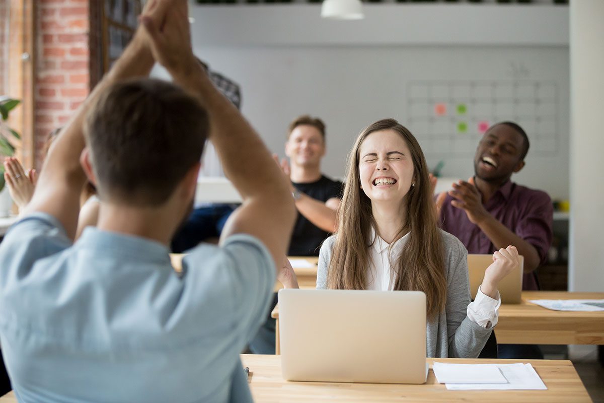 A young woman in an open office setting looking very happy and the other employees are cheering.