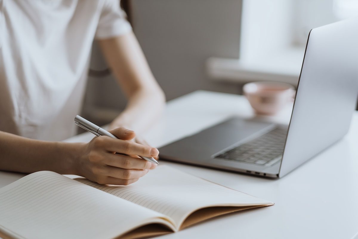 A cropped photo of a person sitting at a desk with a laptop and notebook with a pen in their hand.