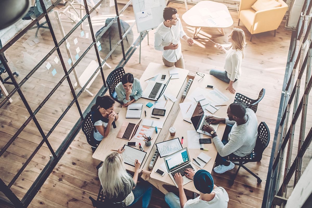 A group of people at a large desk in a casual office setting planning a project with laptops and spreadsheets covering the desk.